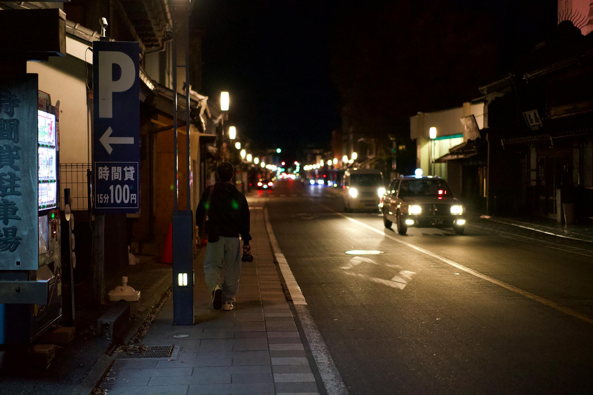 City street at night with a lone pedestrian and vending machine lighting the sidewalk.