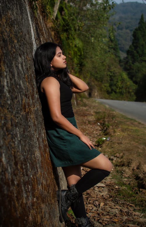 Woman in Black Sleeveless Top and Dark Green Skirt Leaning on a Wall