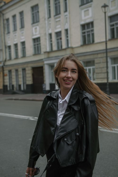 Free A woman with long brown hair, sporting a black leather jacket, smiles while walking in urban streets. Stock Photo