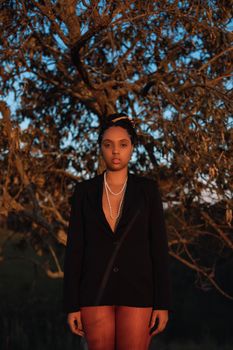 Stylish woman posing in nature with a black blazer and pearl necklace under a tree.