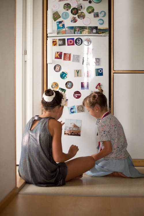 Girls Sitting by a Fridge and Looking at Magnets 