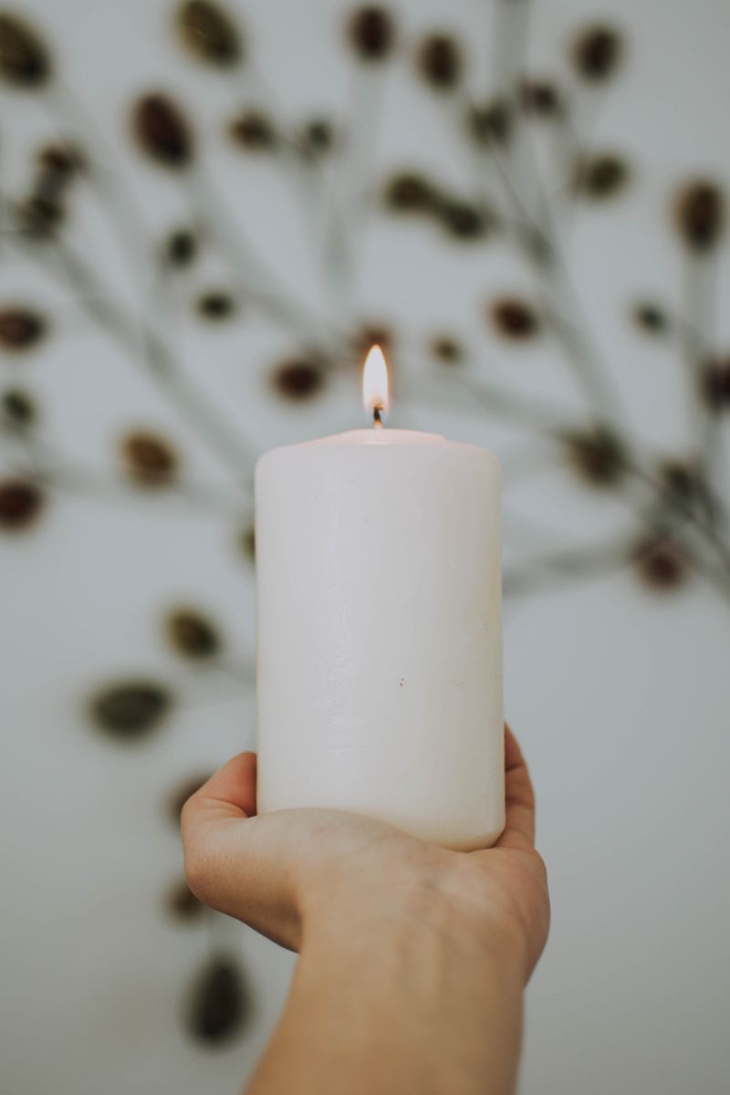 A hand holding a white pillar candle against a pale sky