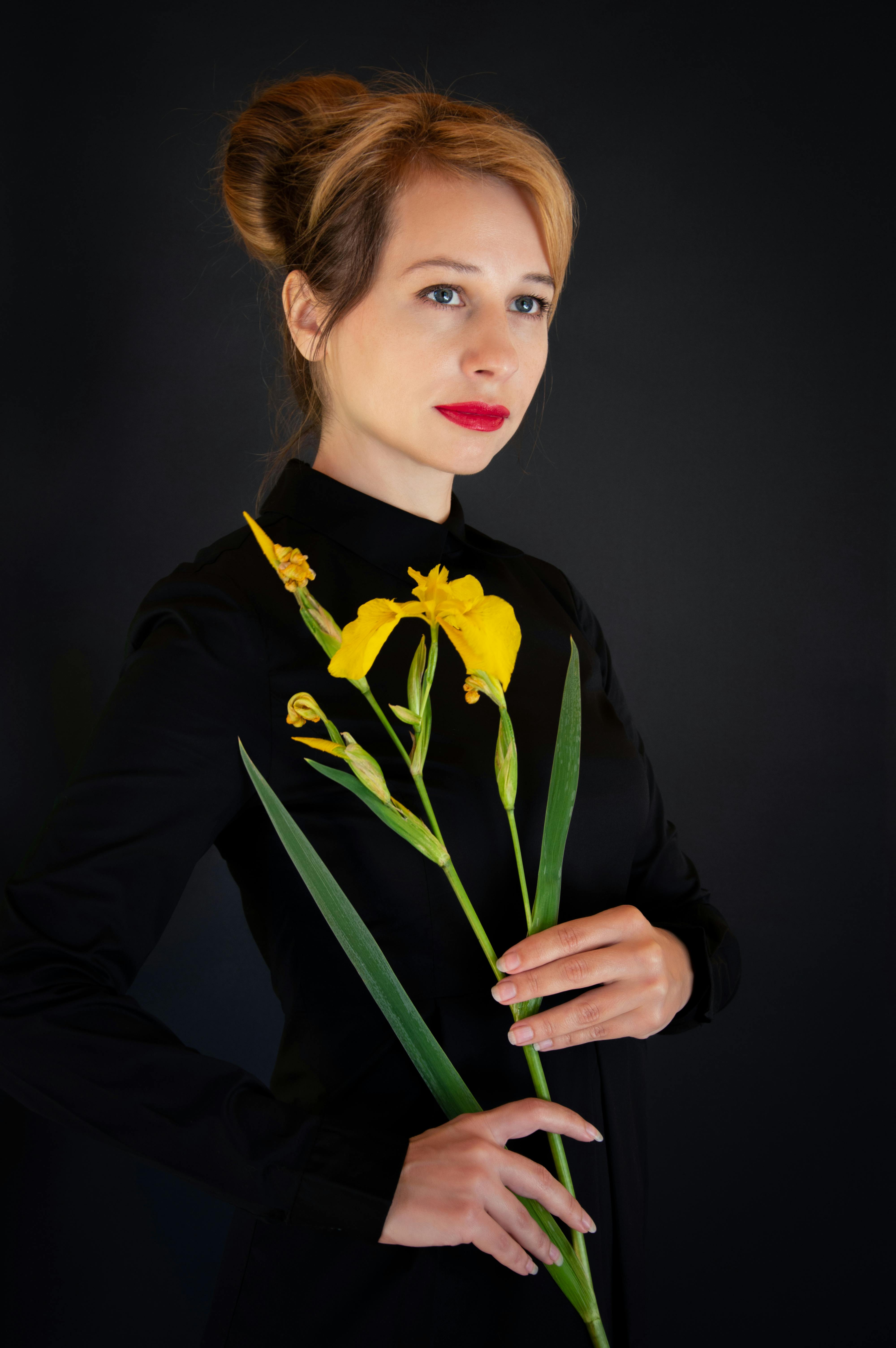 Studio portrait of a blonde woman in black holding a yellow iris, set against a dark background.