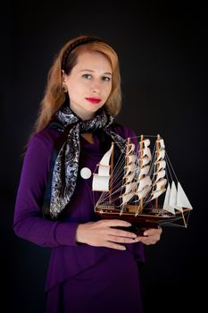 A sophisticated woman in purple attire holding a ship model, set against a black backdrop.