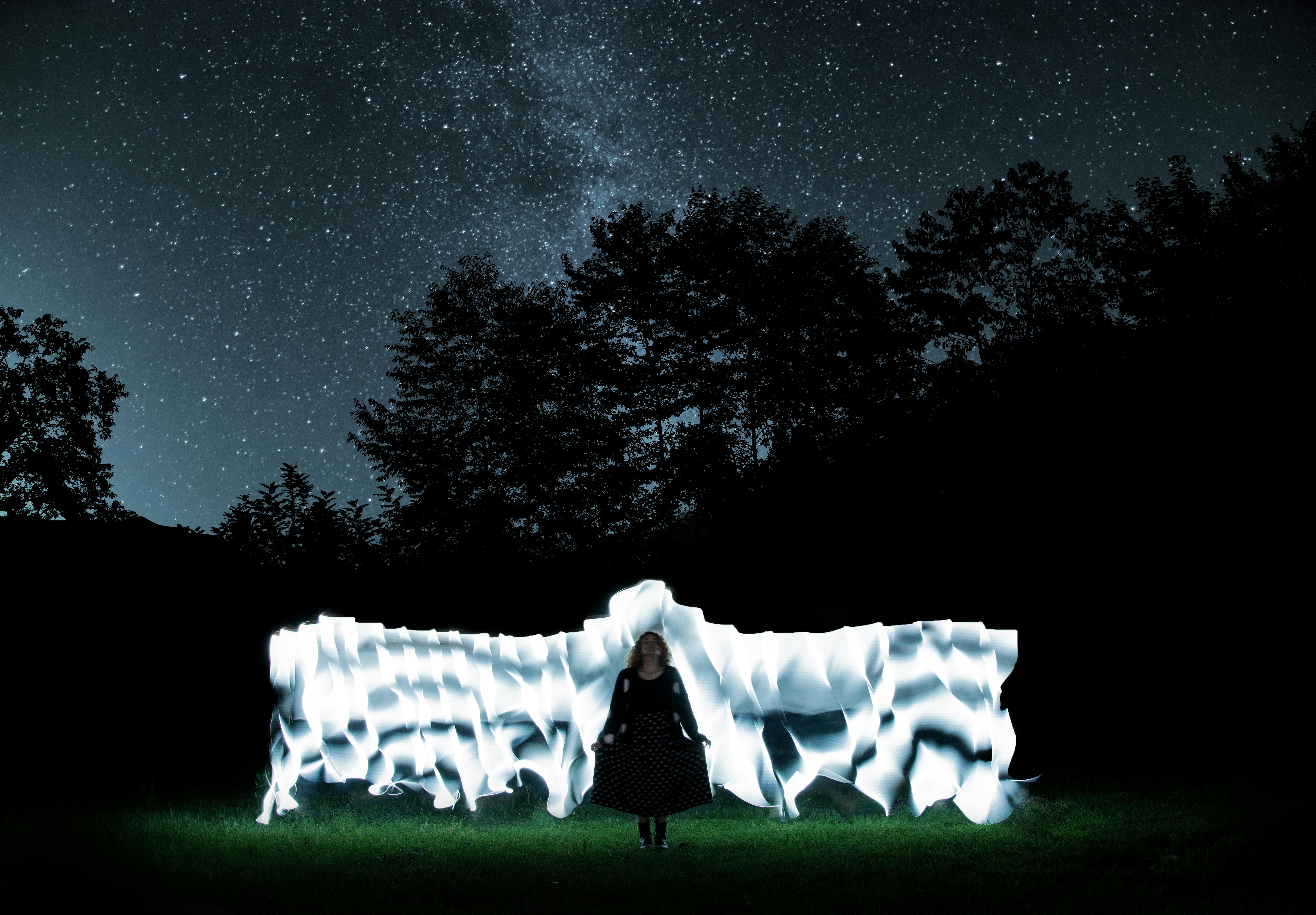 A woman stands under a star-lit night sky, surrounded by artistic light trails.