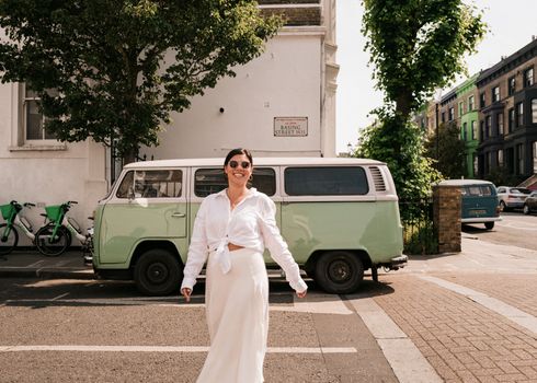 Smiling woman in white outfit enjoying a sunny day near a vintage VW camper in London.