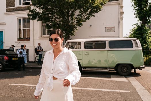 A smiling woman in sunglasses and a white shirt walks confidently on a sunny London street.