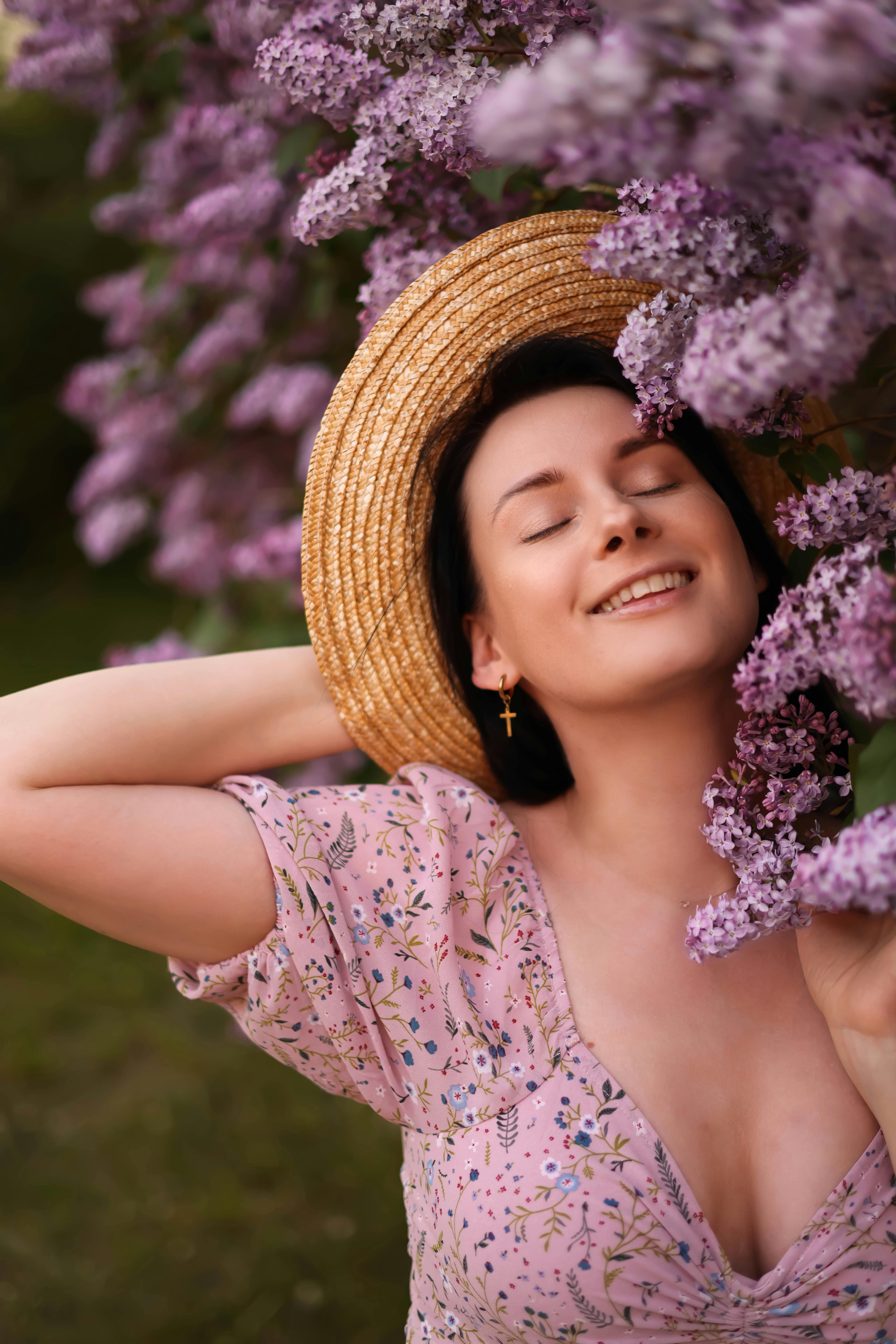 Young woman with hat smelling lilac flowers, eyes closed, in a tranquil spring setting.