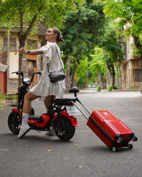 Stylish woman on scooter towing a red suitcase in a tree-lined Istanbul street.