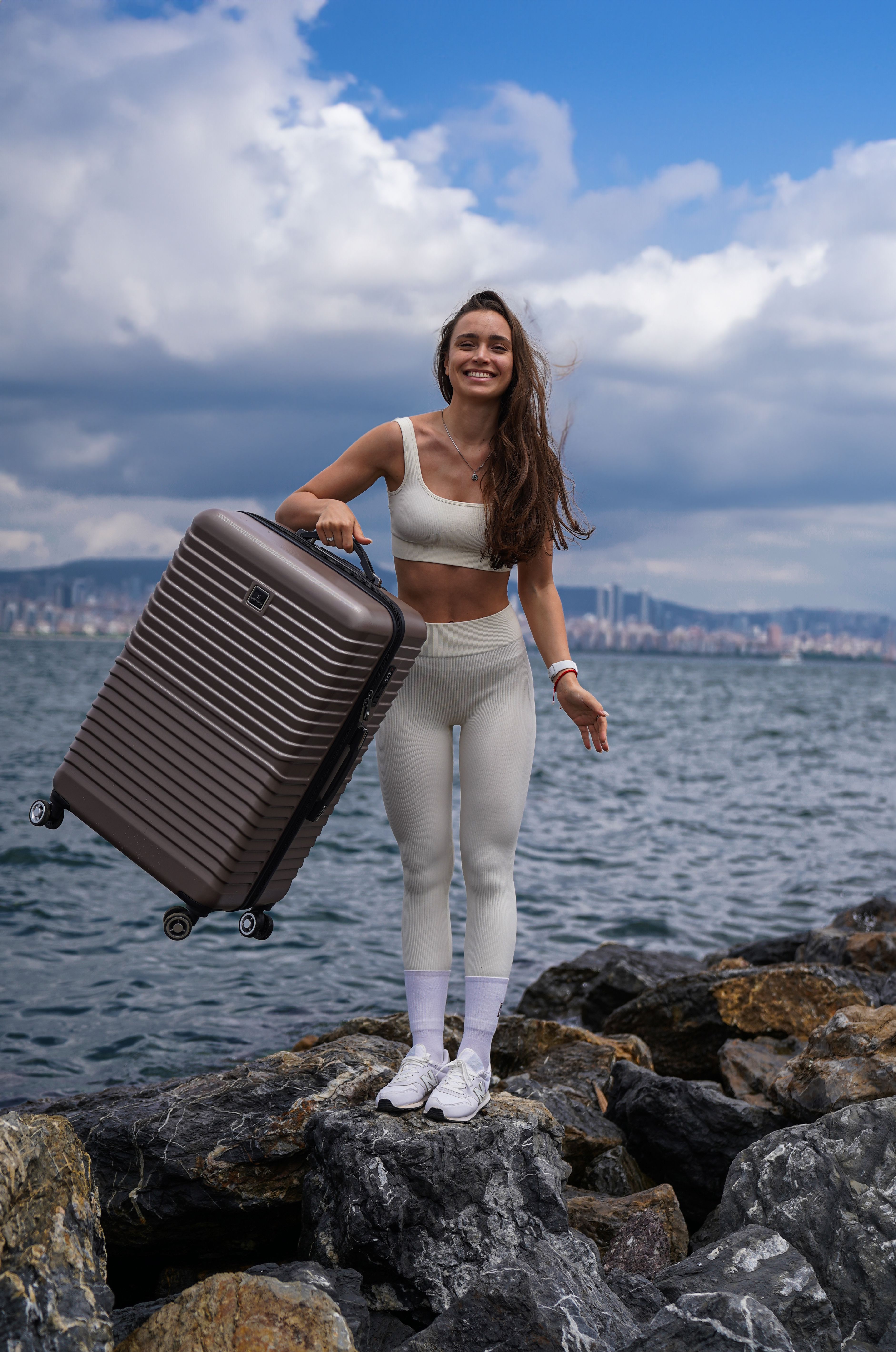 Smiling woman in sportswear holding luggage on rocky Istanbul seaside, enjoying leisure outdoors.