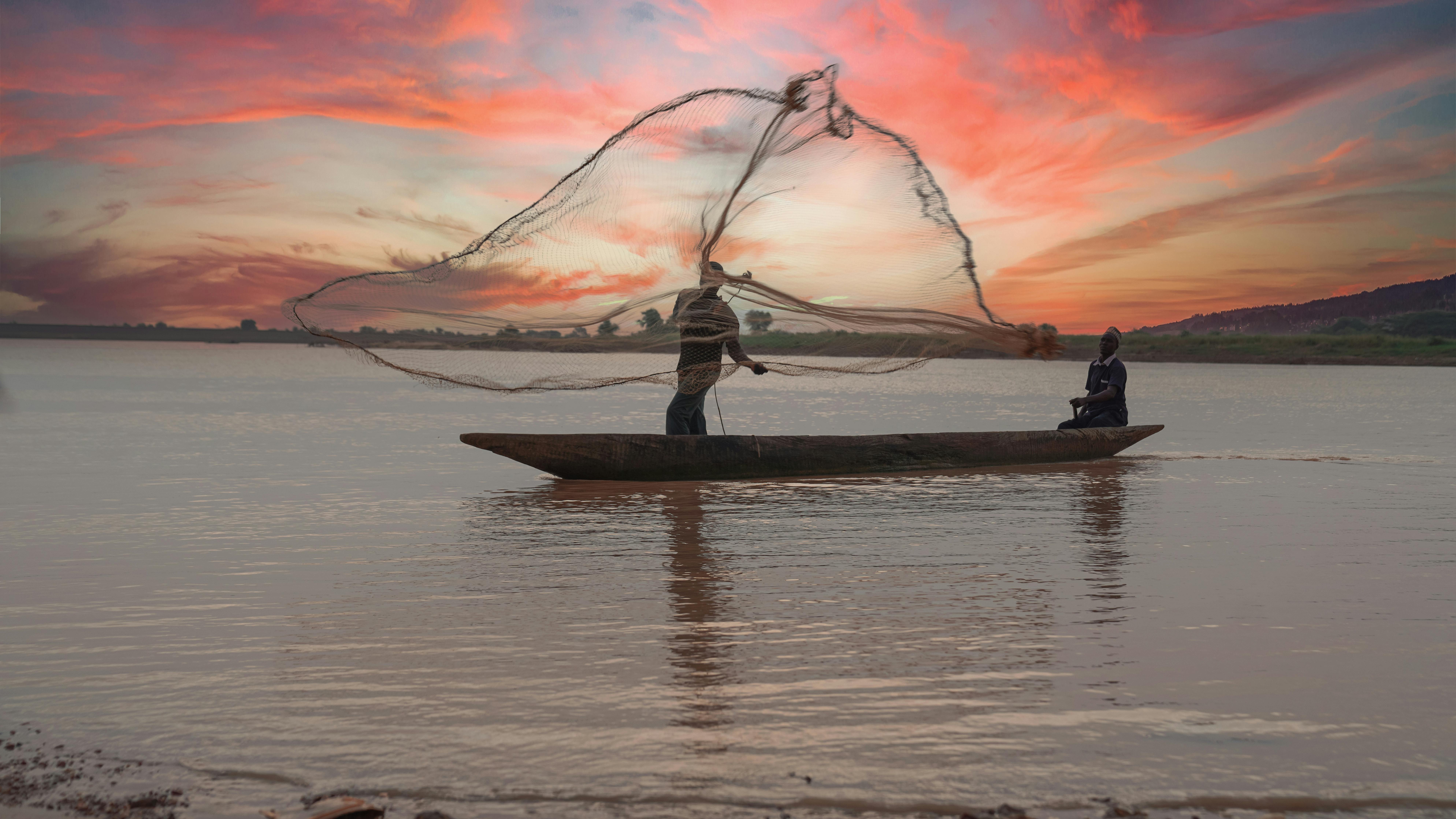 Two fishermen casting nets from a boat on a calm river during a serene sunset in Calabria, Italy.