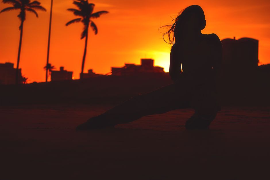 A silhouette of a woman stretching outdoors against a vibrant sunset with palm trees in the background.