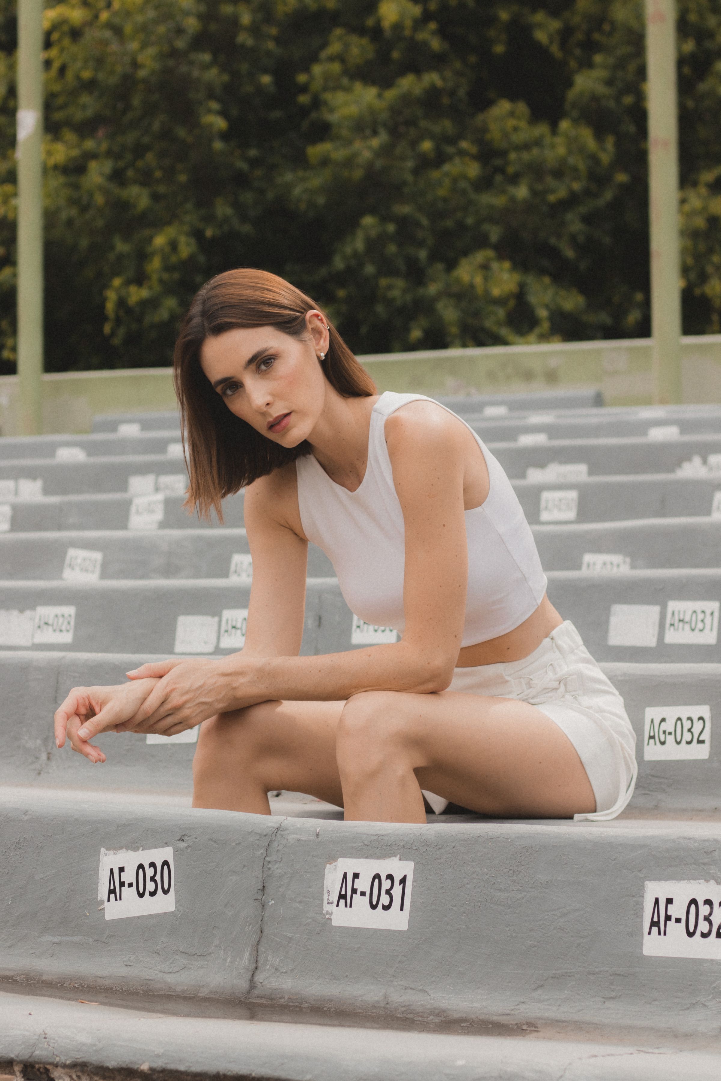 Confident woman in white crop top and shorts seated in outdoor stadium stands.