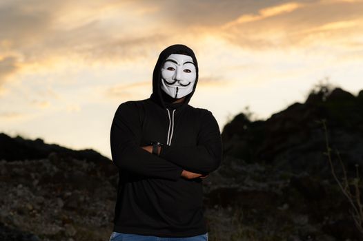 A man wearing a Guy Fawkes mask and hoodie stands outdoors at sunset, creating a mysterious silhouette.