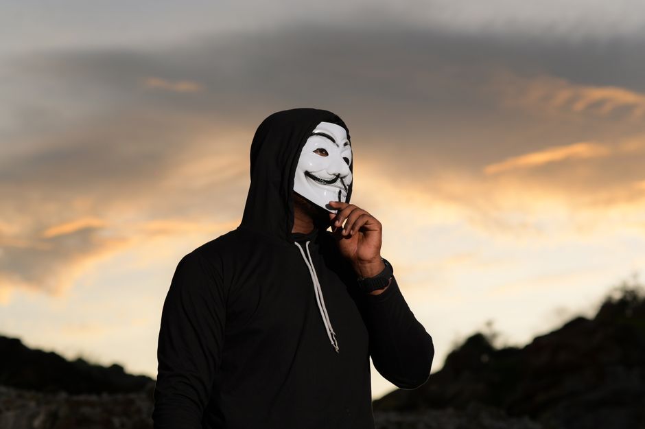 Anonymous individual wearing a Guy Fawkes mask standing outdoors during a stunning sunset.
