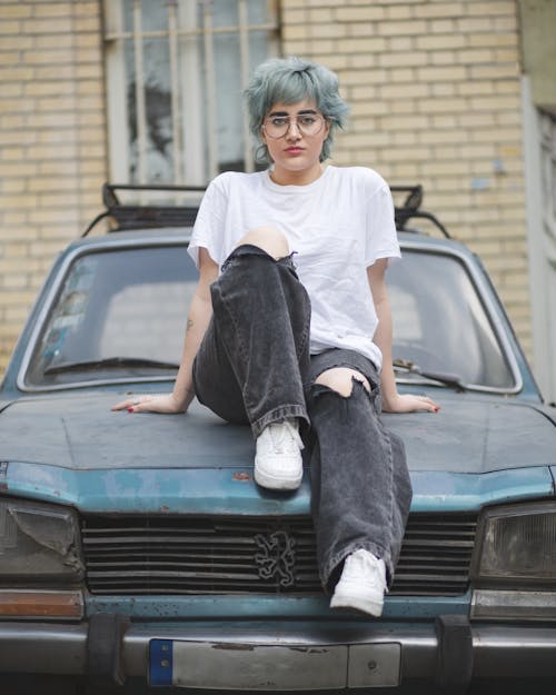 Blue-haired Model in a White T-shirt and Distressed Jeans Posing on the Hood of an Abandoned Blue Car