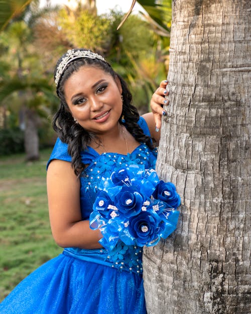 Model in a Blue Evening Dress and Tiara Posing by a Tree in the Park