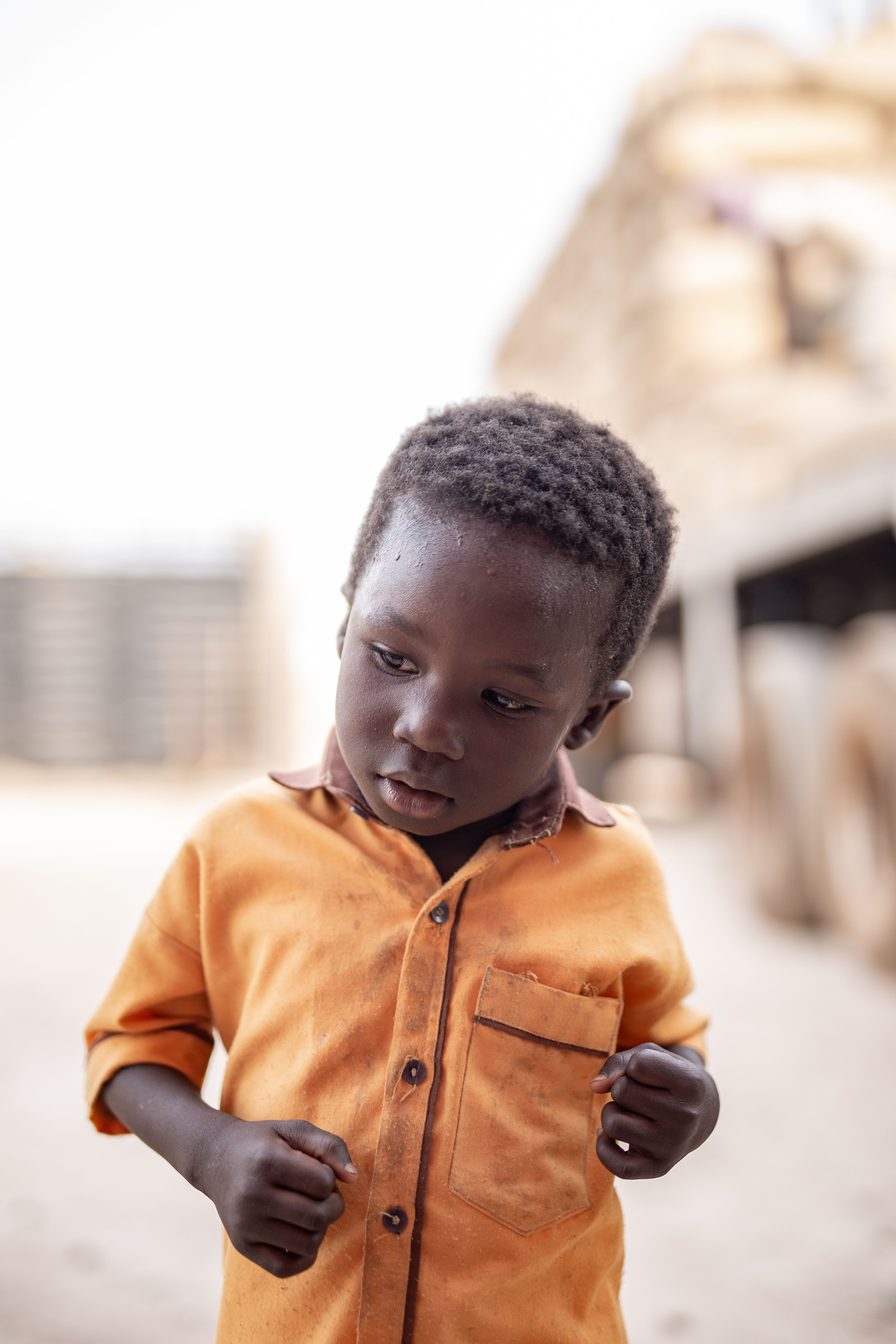 A young boy in an orange shirt wandering outdoors in Sampa, Bono Region, Ghana.