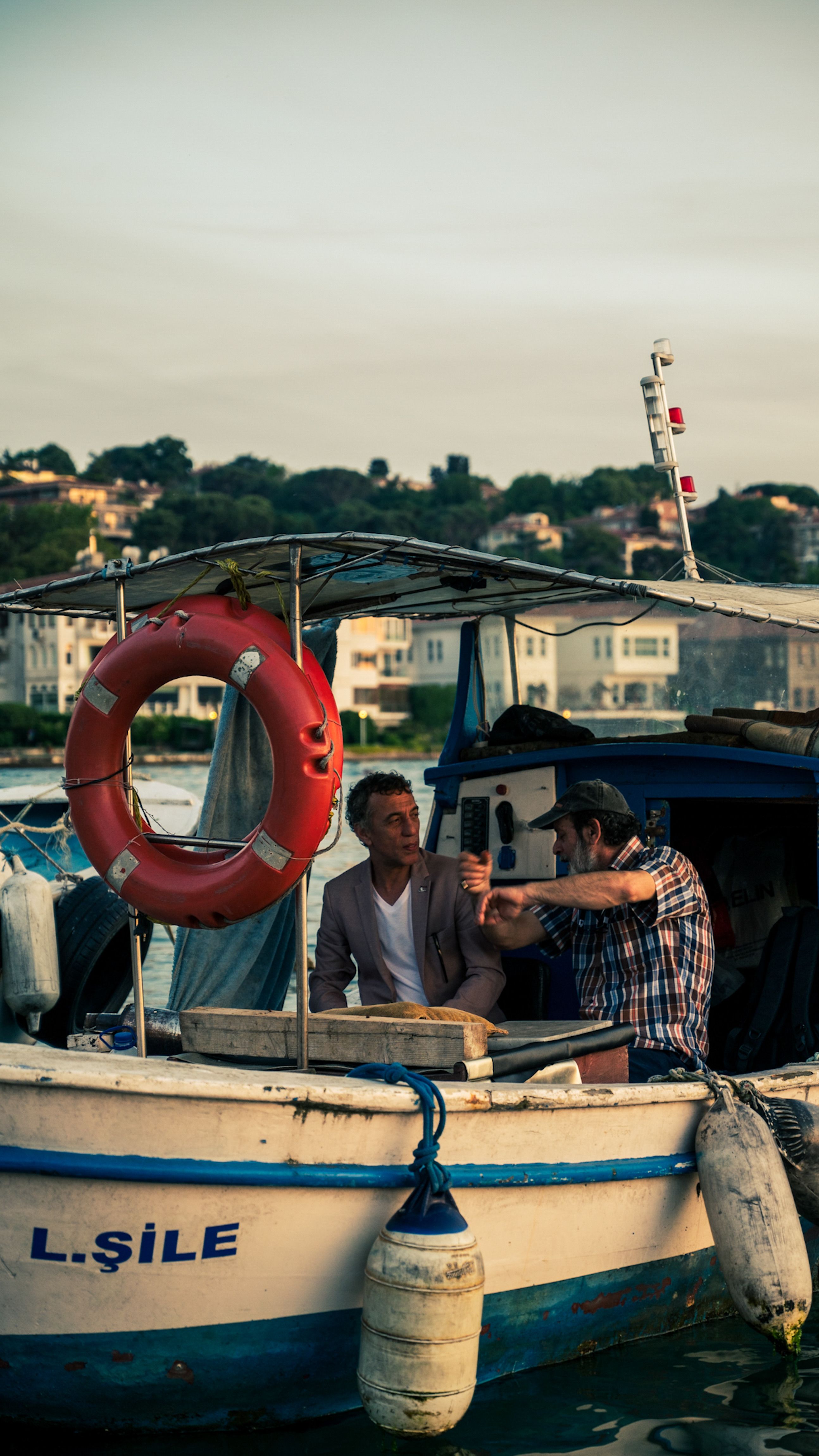 Two men engaged in conversation on a fishing boat with a lifebuoy, set against a picturesque coastal town.