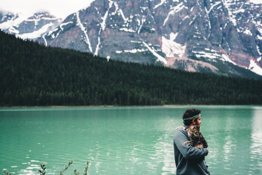 A man holding a cat by a stunning lake in Jasper, Alberta, Canada with mountain backdrop.