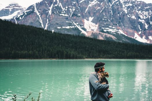 Man holds fluffy cat by emerald lake with stunning mountain backdrop in Jasper National Park.
