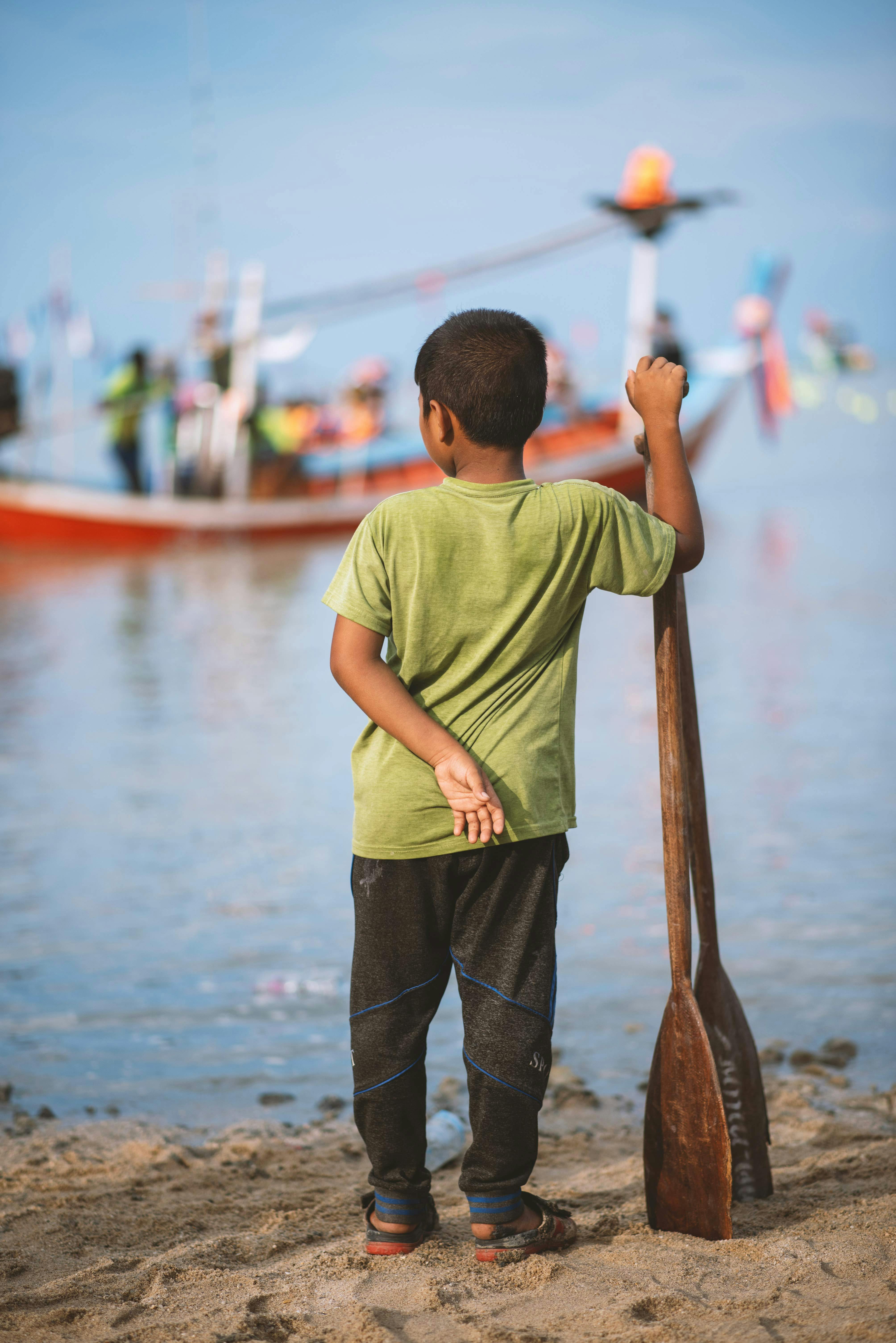 A boy in a green shirt stands on the beach holding a paddle, looking at a boat.