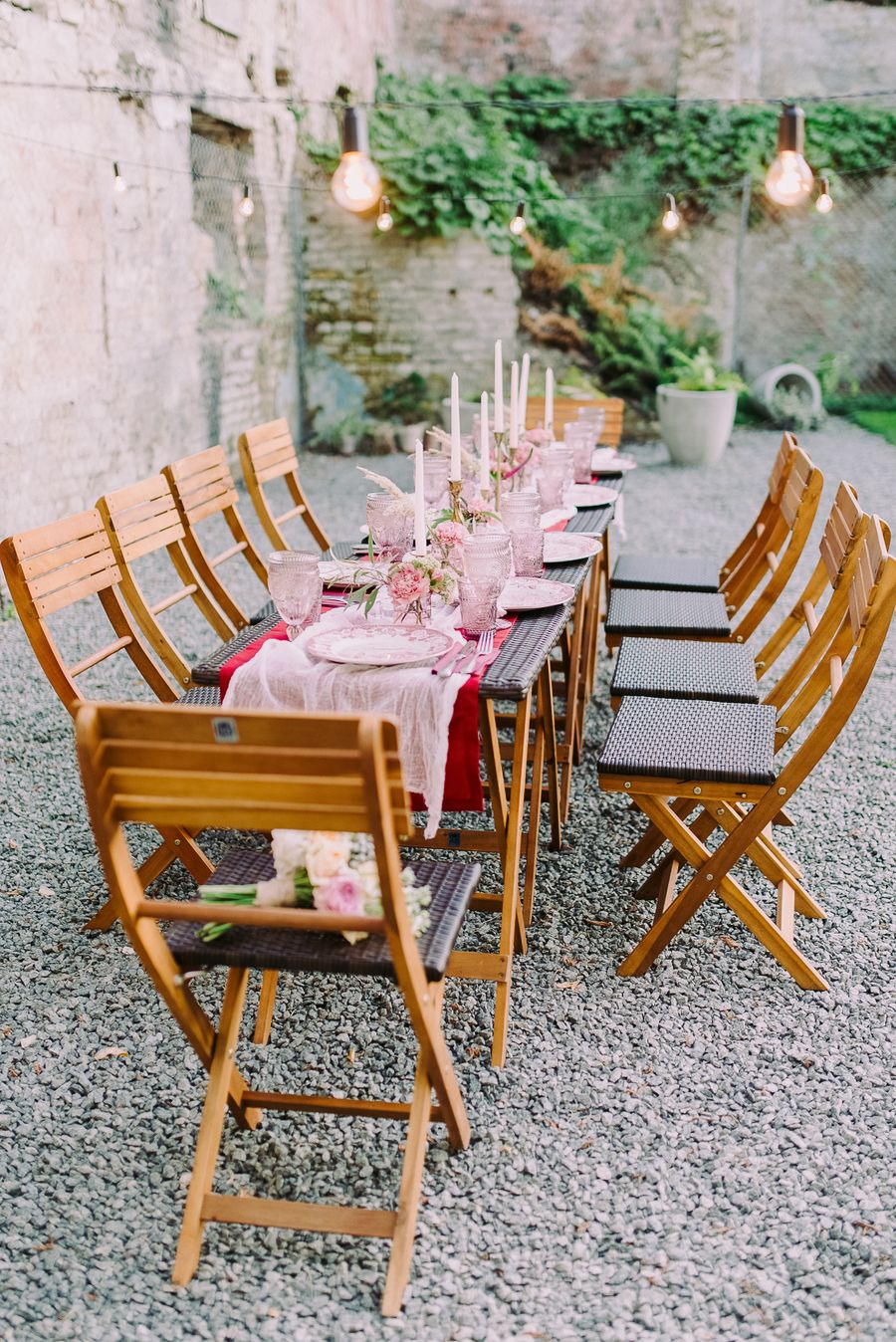 A long dinner table laid with a pink cloth, candles and wooden folding chairs in a stone courtyard