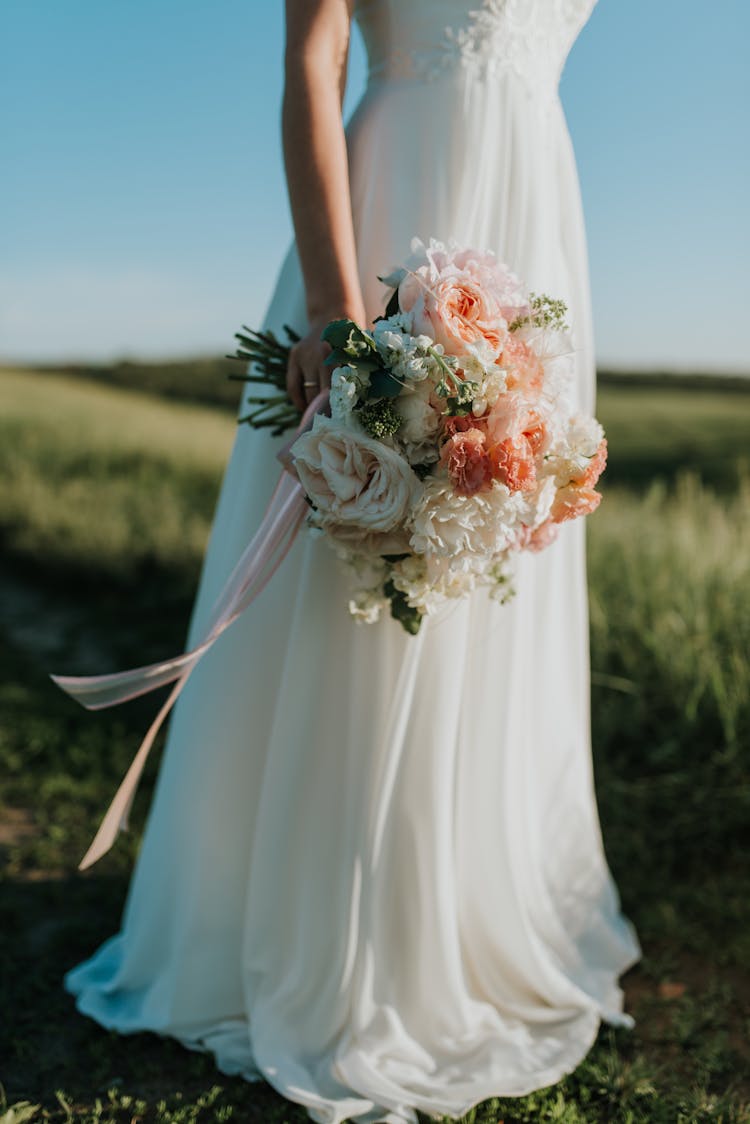 Woman Wearing White Wedding Dress Holding Flower Bouquet Standing On Green Field