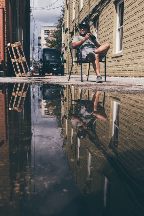 Free Man sitting in an alleyway, reflected in a puddle on a sunny day. Stock Photo