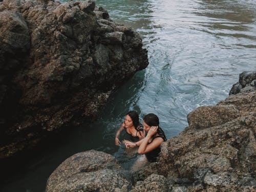 Two Women Swimming on Body of Water Beside Rocky Cliff