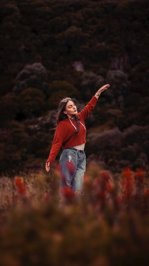 Free A young woman in casual red clothing poses gracefully in a vibrant meadow at sunset. Stock Photo