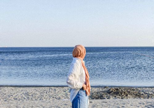 Free A woman in a hijab stands on the beach, gazing at the tranquil sea under a clear sky. Stock Photo
