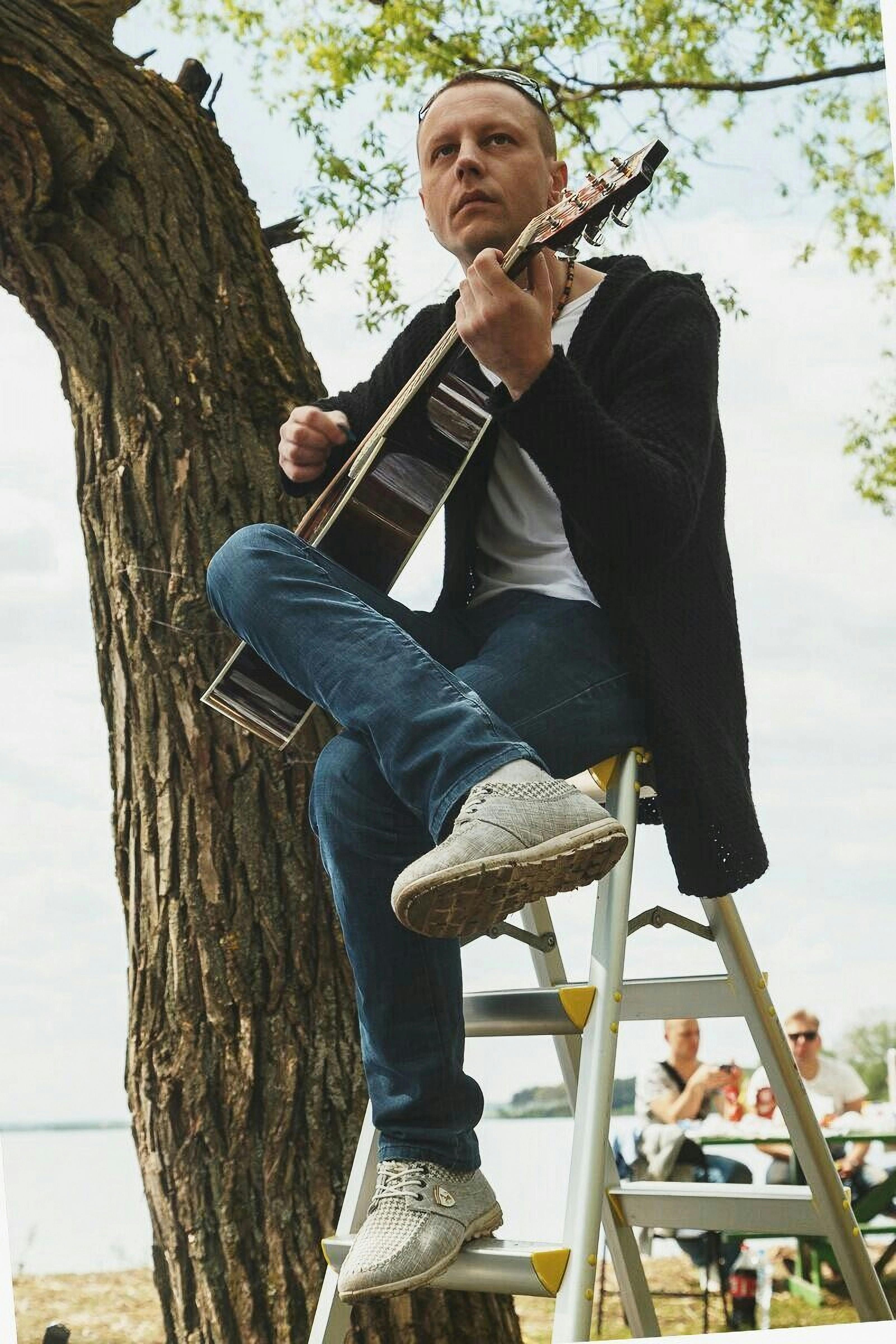 A man plays an acoustic guitar while sitting on a ladder under a tree in the park.