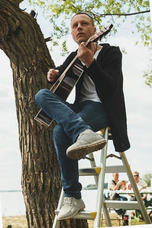Man Sitting Outside on a Ladder and Playing the Guitar 