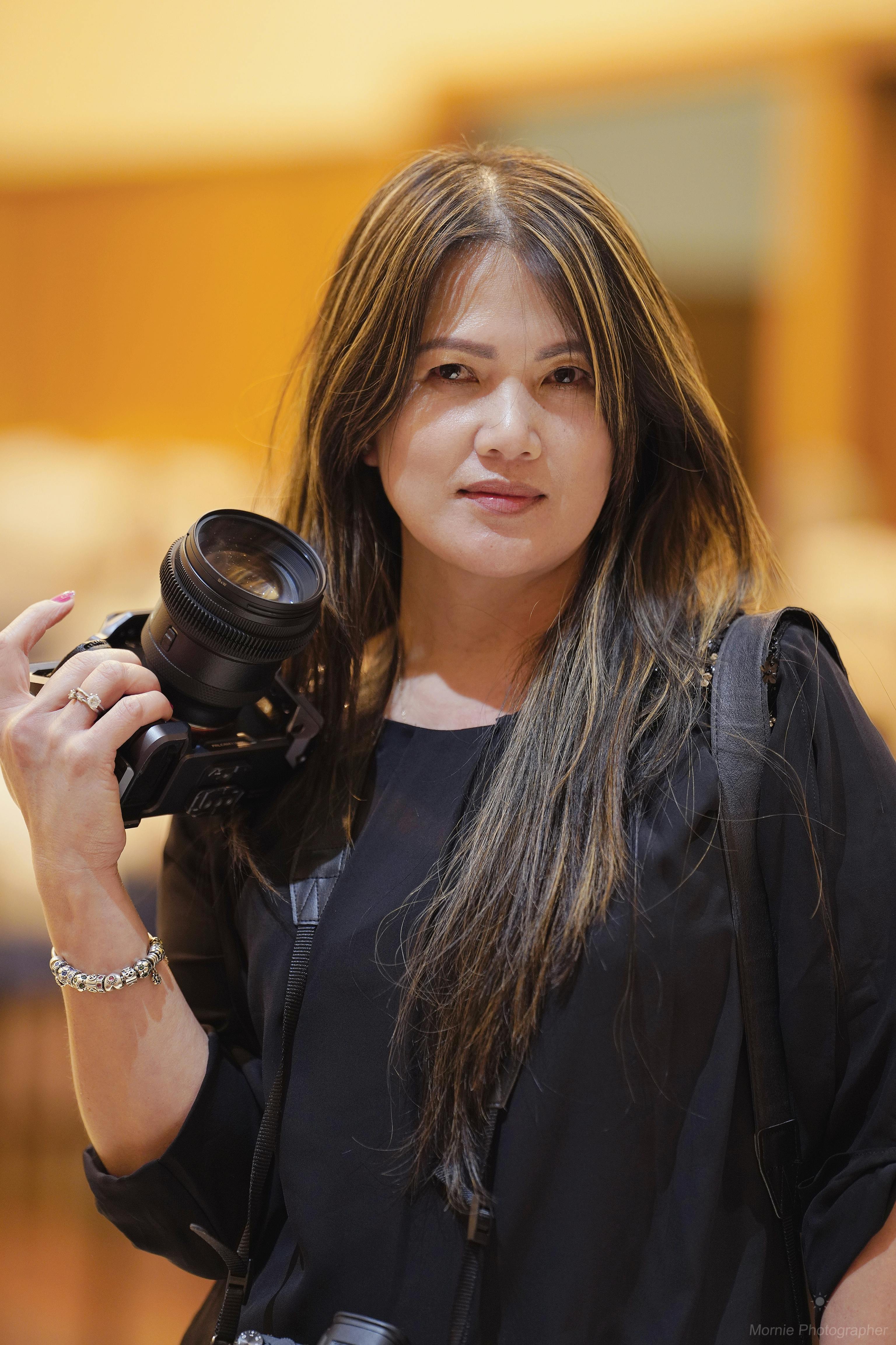 Asian female photographer holding a camera indoors, wearing a black blouse and bracelet.
