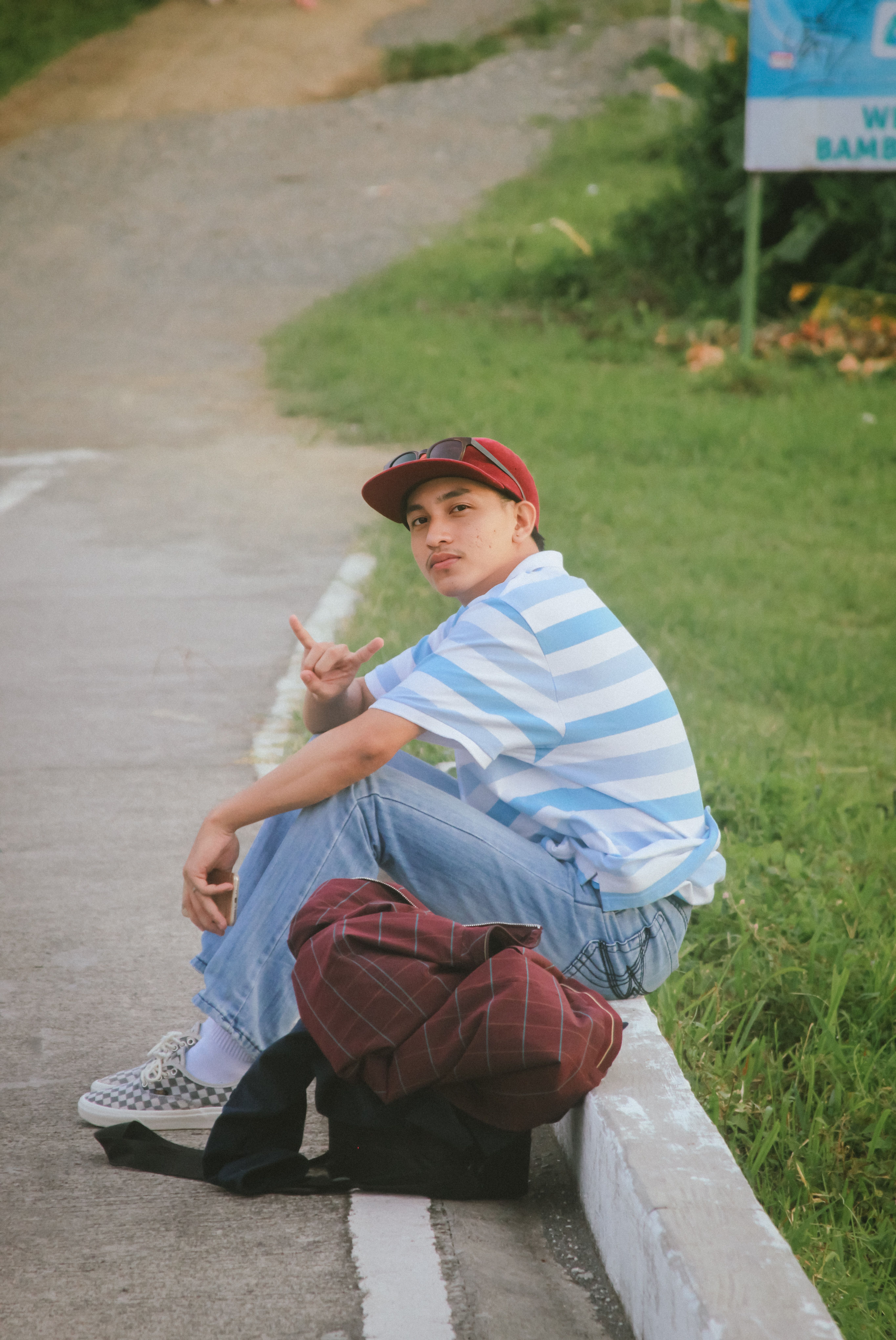 A young man in a striped shirt and cap sits on the sidewalk posing with a casual demeanor.