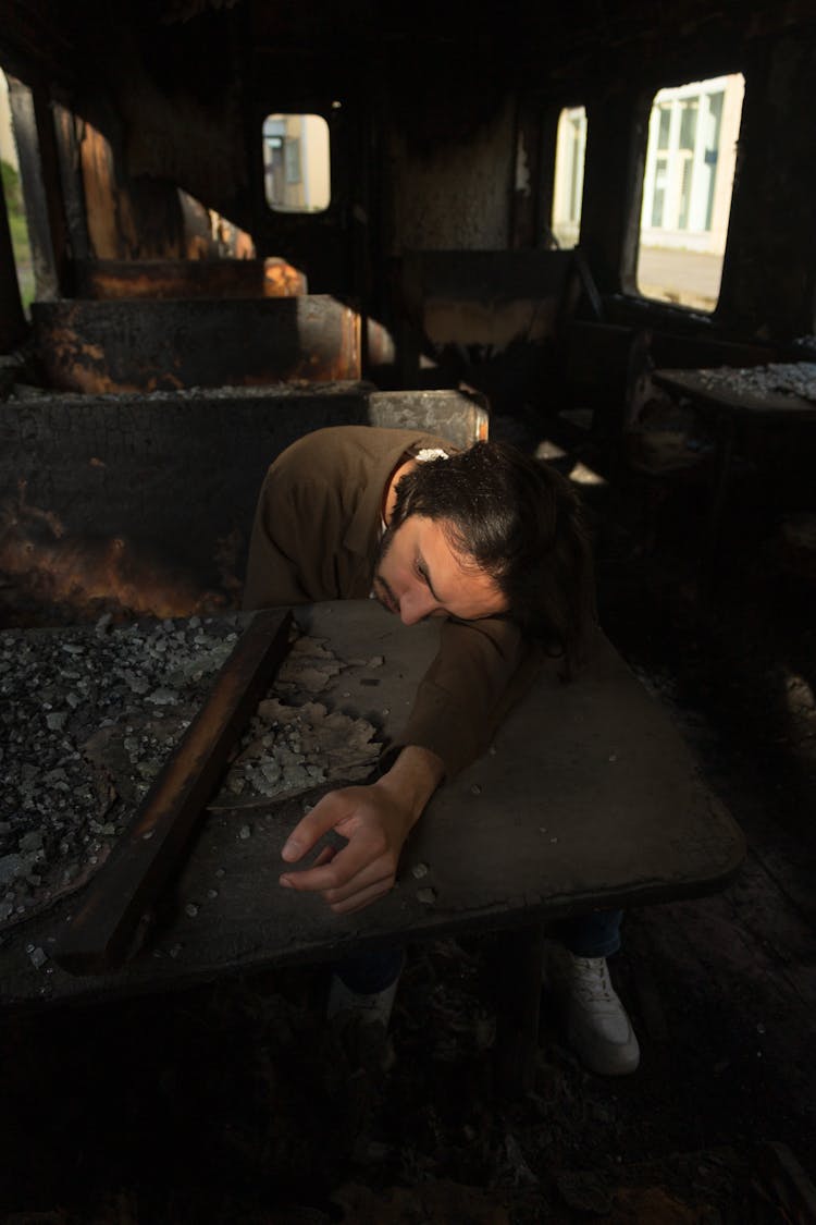 A Man Sitting And Resting His Head On A Table In An Abandoned, Burnt Down Vehicle 