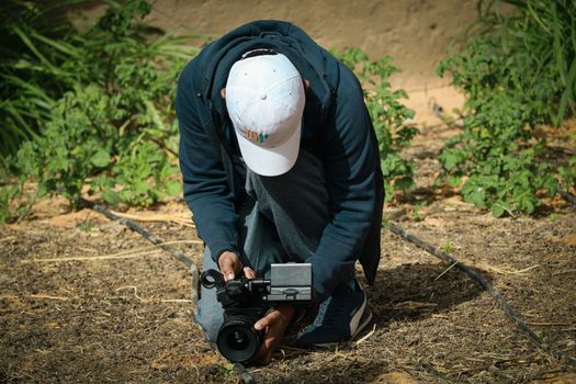 A person in a white cap operates a camera in an outdoor garden scene.