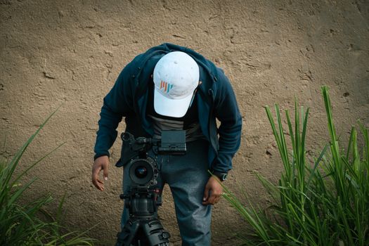 A man adjusts a camera setup outdoors near a grassy wall.
