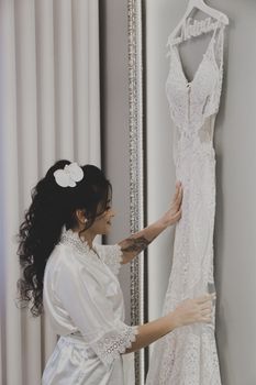 Bride touching lace wedding dress hanging on wall, showcasing intricate details.