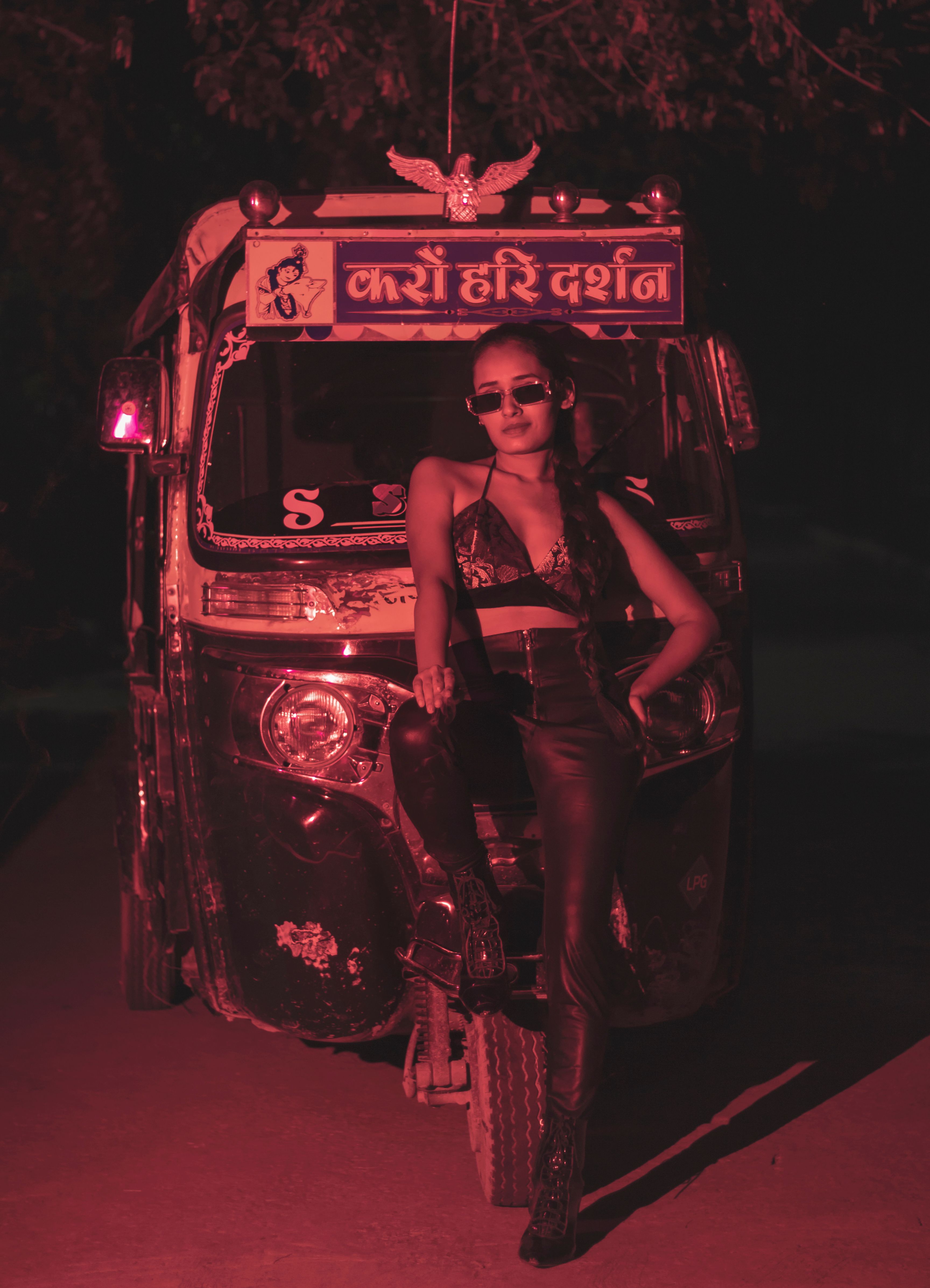 Trendy woman in sunglasses posing against a vibrant pink-lit auto rickshaw at night.