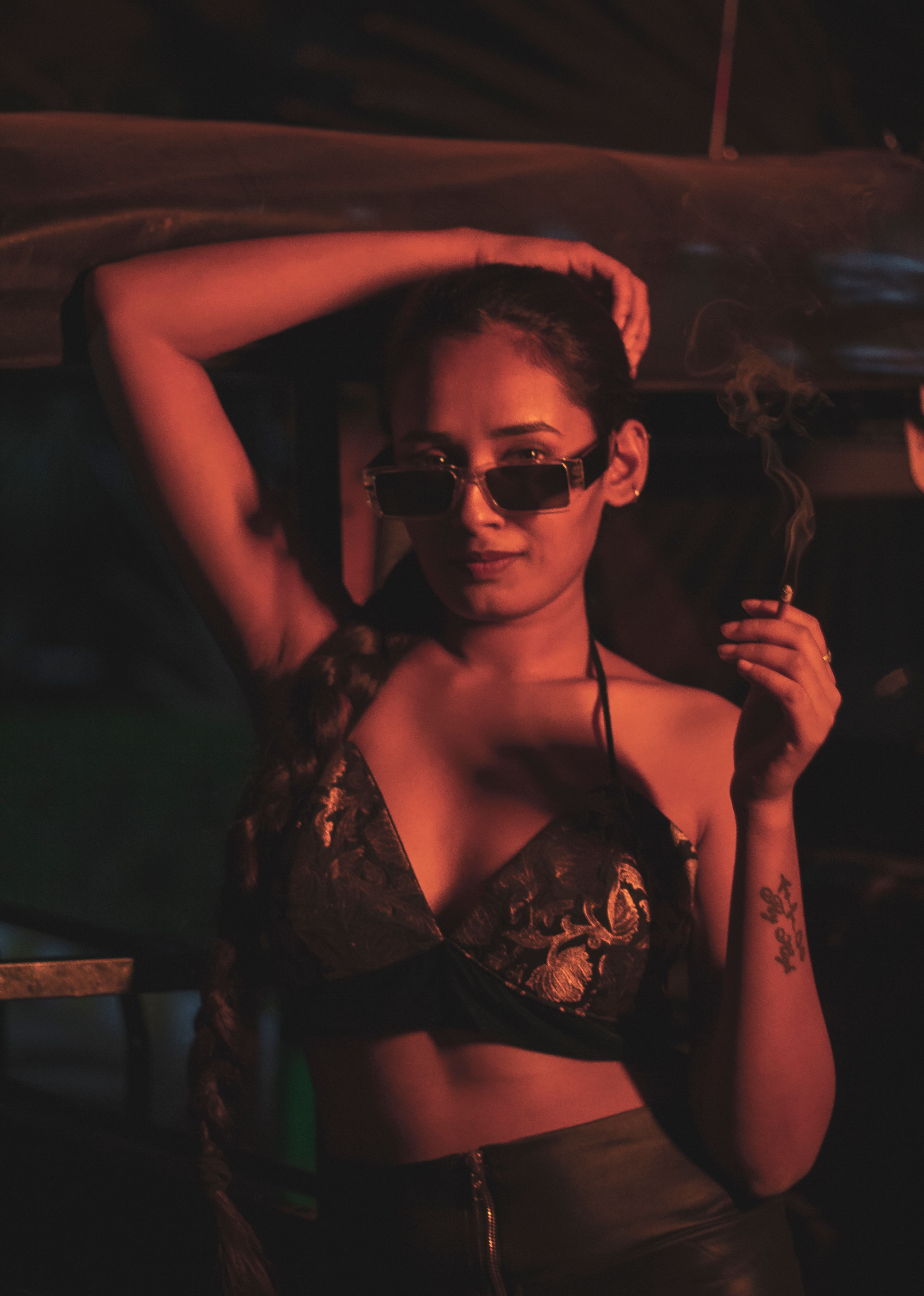 Young woman with sunglasses smoking, bathed in moody red lighting, posing confidently.