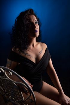 Moody studio shot of a tattooed woman in a black bodysuit sitting on a chair against a blue background.