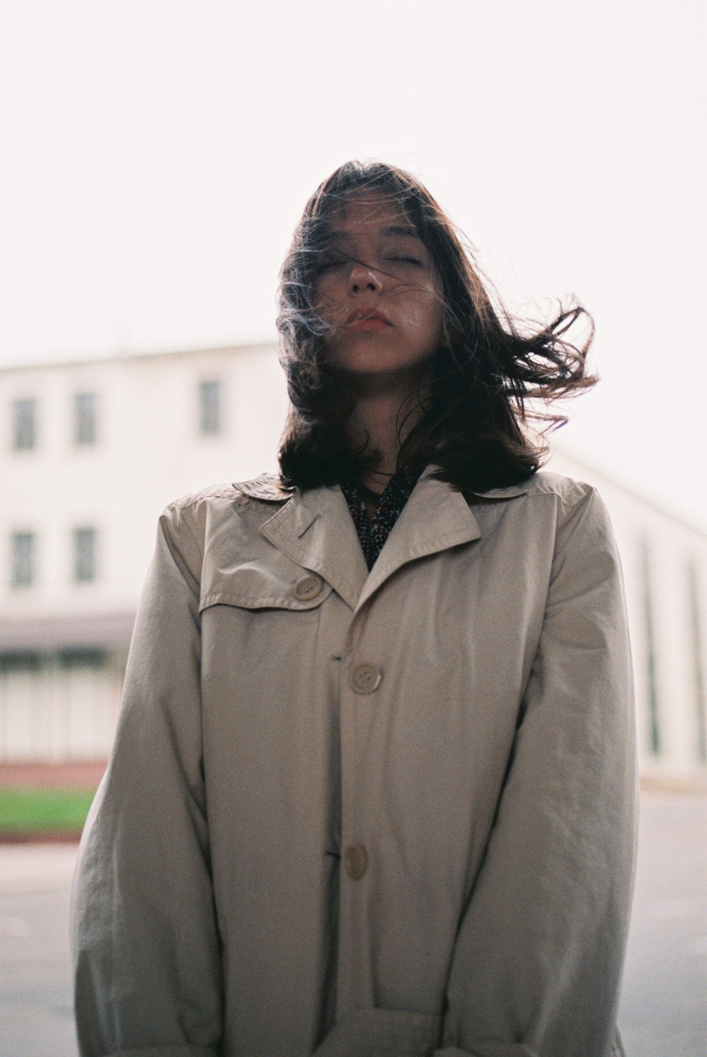 Stylish young woman wearing a trench coat, posing dramatically in a city setting with wind-swept hair.