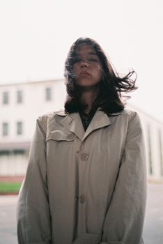 Stylish young woman wearing a trench coat, posing dramatically in a city setting with wind-swept hair.