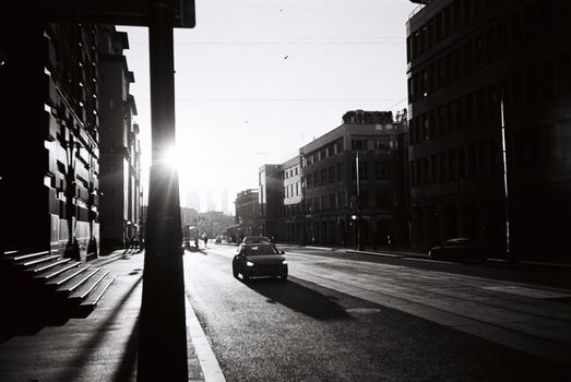 Monochrome view of a street with buildings and cars during sunset, creating stark shadows.