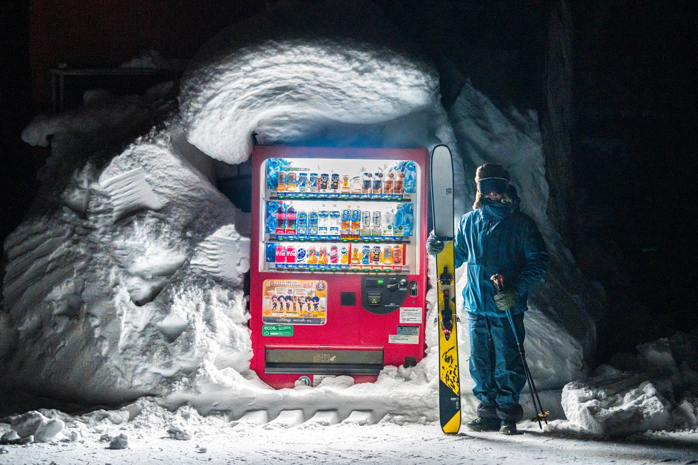 A skier stands by an illuminated vending machine in snowy Hokkaido, Japan at night.