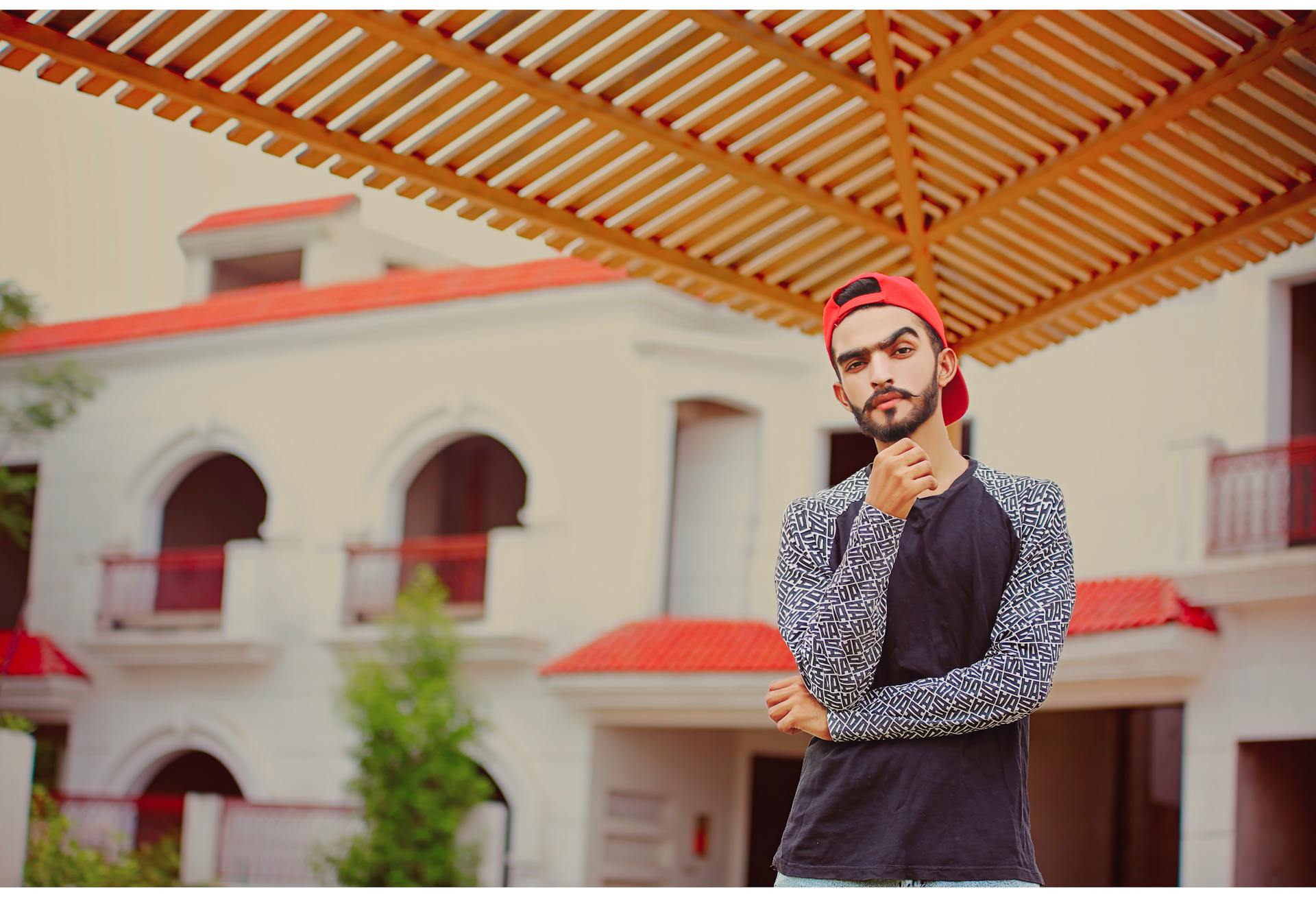 Young man in casual outfit posing outdoors under a pergola with red-tiled building in the background.