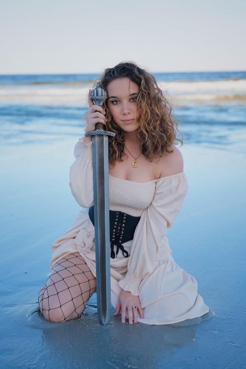 Free A woman in medieval attire kneels on the shore, holding a sword, embodying a historical fantasy theme. Stock Photo