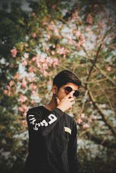 一名年轻人在户外盛开的树旁戴着太阳镜摆姿势，拍摄时尚照片